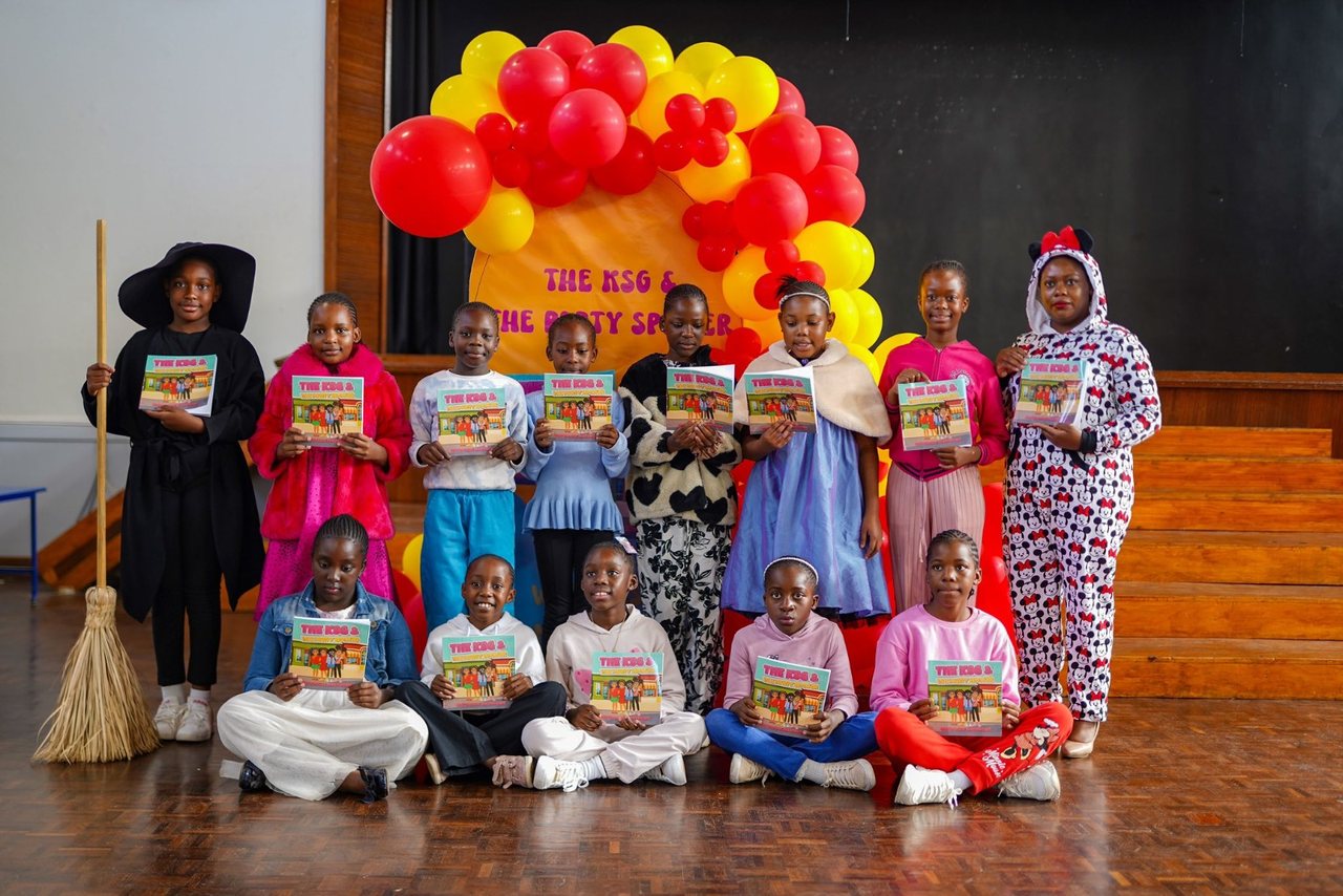 Pupils in costume holding copies of The KSG & The Party Speaker at its school launch, beneath a red and yellow balloon arch.